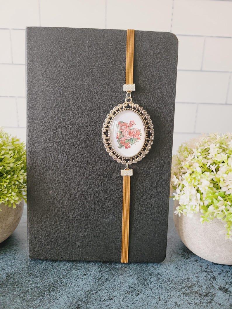 May include: A black notebook featuring a vertical mustard-colored elastic band. Attached to the band is an oval-shaped silver-toned metal pendant with a decorative filigree border, framing a white insert with a pink and green floral illustration.