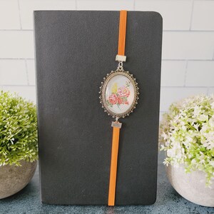 May include: A black notebook featuring a vertical orange elastic band. Attached to the band is an oval-shaped silver-toned metal pendant with a decorative filigree border, containing a floral illustration of pink and yellow flowers on a white background.