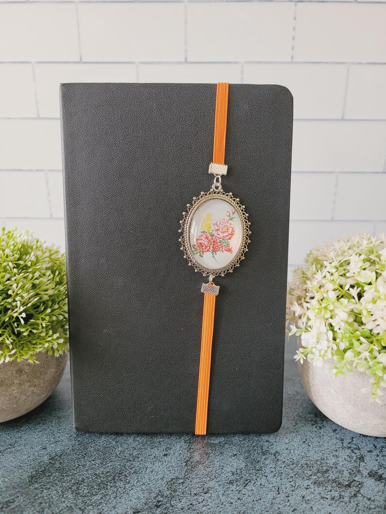 May include: A black notebook featuring a vertical orange elastic band. Attached to the band is an oval-shaped silver-toned metal pendant with a decorative filigree border, containing a floral illustration of pink and yellow flowers on a white background.