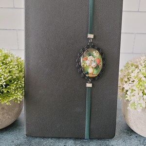 May include: A dark gray notebook featuring a vertical dark green elastic band. Attached to the band is an oval-shaped black filigree frame containing a miniature print of a colorful floral bouquet in a vase, secured with small silver-toned metal accents.