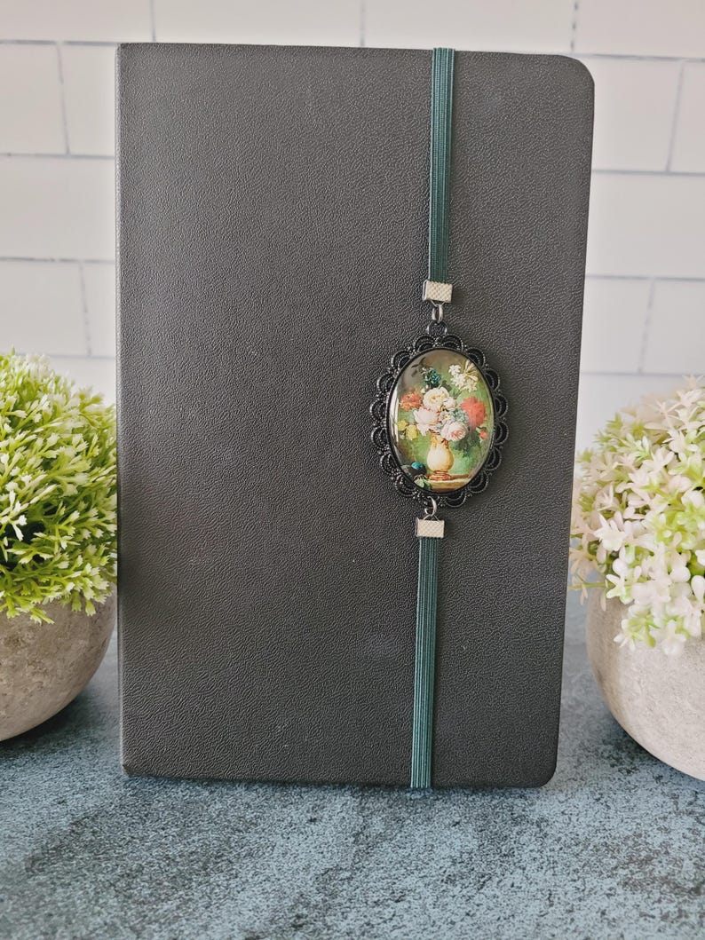 May include: A dark gray notebook featuring a vertical dark green elastic band. Attached to the band is an oval-shaped black filigree frame containing a miniature print of a colorful floral bouquet in a vase, secured with small silver-toned metal accents.