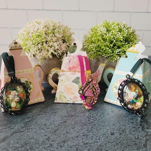 May include: Three decorative teapot-shaped boxes in pink, floral, and light blue, each holding an accessory. Two feature oval black frames with floral art, while the center one displays a pink butterfly brooch with embedded crystals.