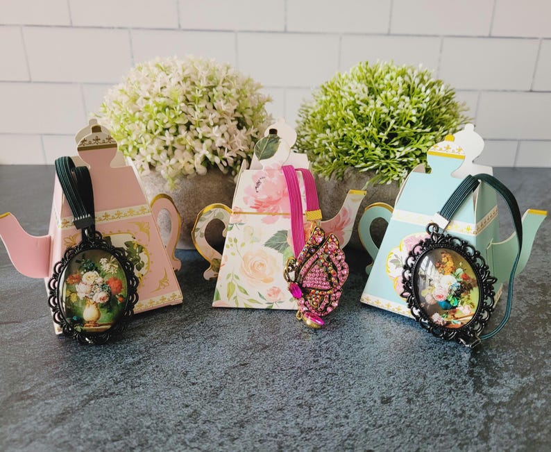 May include: Three decorative teapot-shaped boxes in pink, floral, and light blue, each holding an accessory. Two feature oval black frames with floral art, while the center one displays a pink butterfly brooch with embedded crystals.
