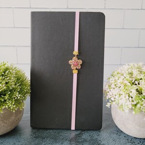 May include: A black notebook featuring a vertical light pink elastic band closure. Centered on the band is a gold-toned metal flower charm with pink crystal accents, held in place by two small gold-toned metal rectangular sliders.