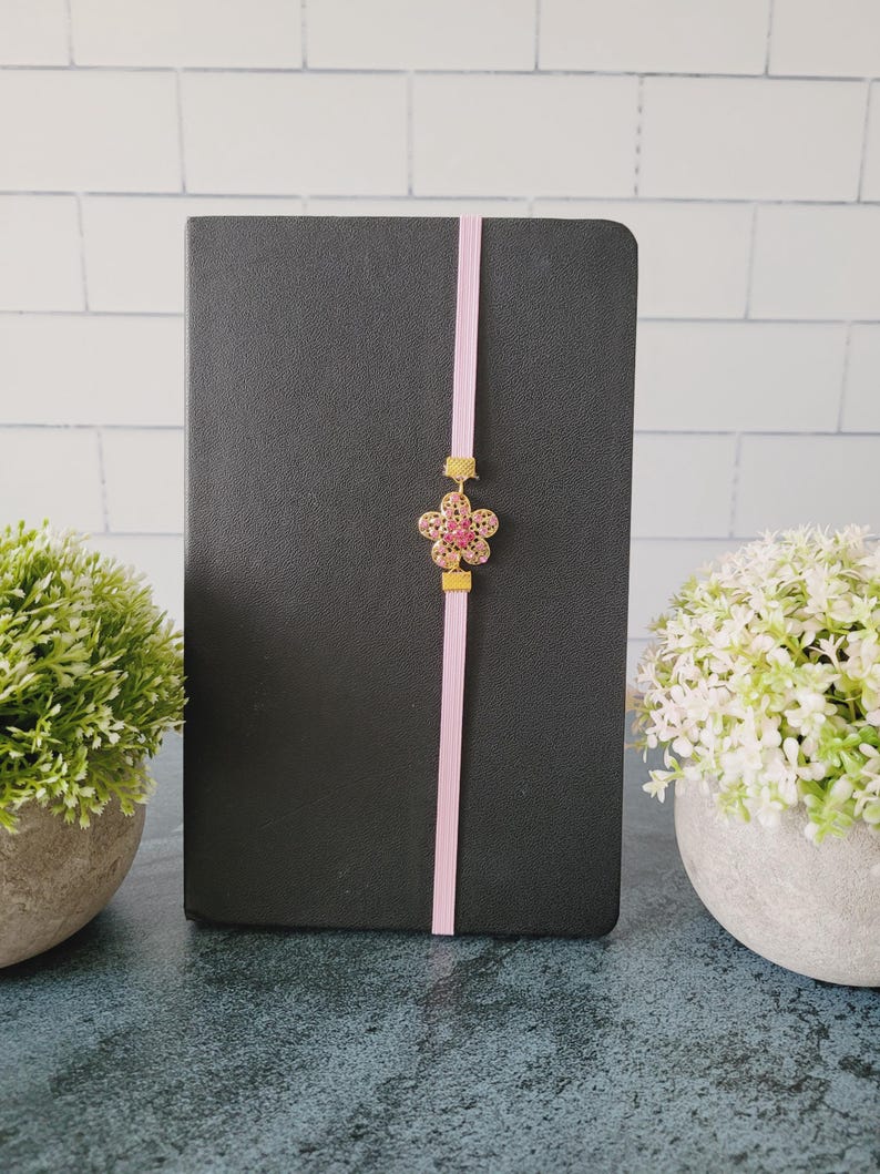May include: A black notebook featuring a vertical light pink elastic band closure. Centered on the band is a gold-toned metal flower charm with pink crystal accents, held in place by two small gold-toned metal rectangular sliders.