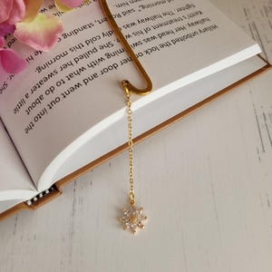 May include: A gold bookmark with a flower-shaped charm. The bookmark is shaped like a letter L and has a chain attached to the charm.