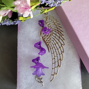 Könnte beinhalten: Ein silberner Flügel-Anhänger mit einer lila Blume und einem Feendesign. Der Anhänger befindet sich in einer rosa Geschenkbox.