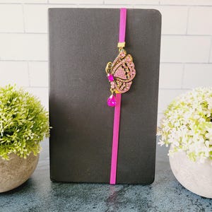 May include: A black notebook featuring a vertical bright pink elastic band. Attached to the band is a decorative gold-toned butterfly ornament encrusted with small pink rhinestones and a single dangling teardrop-shaped pink crystal accent.