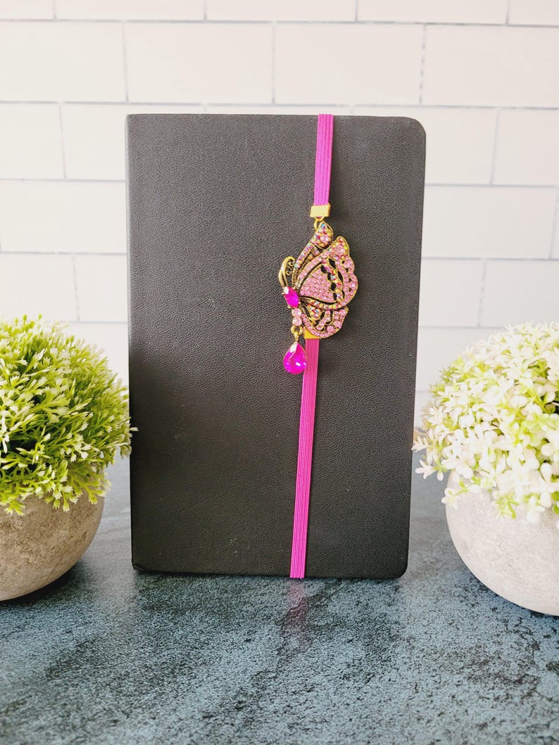 May include: A black notebook featuring a vertical bright pink elastic band. Attached to the band is a decorative gold-toned butterfly ornament encrusted with small pink rhinestones and a single dangling teardrop-shaped pink crystal accent.