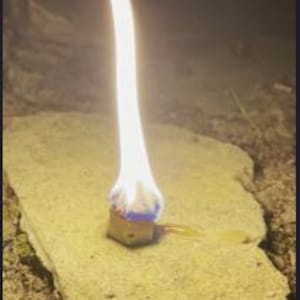 May include: A small, brown, square-shaped fire starter with a blue and red design is lit, producing a tall, bright flame.
