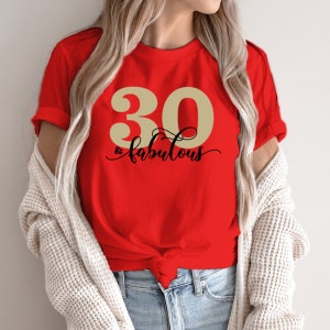 K&ouml;nnte beinhalten: Rotes T-Shirt mit einer goldenen Zahl 30 und dem Text "& fabulous" in schwarzer Schrift.
