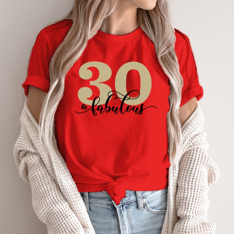 K&ouml;nnte beinhalten: Rotes T-Shirt mit einer goldenen Zahl 30 und dem Text "& fabulous" in schwarzer Schrift.