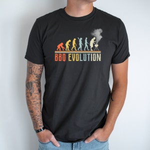 May include: A black t-shirt with a vintage-style graphic of the evolution of grilling. The graphic shows a series of stick figures progressing from a caveman holding a stick over a fire to a modern man grilling on a barbecue. The text "BBQ EVOLUTION" is printed below the graphic.