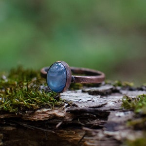 May include: A copper ring with a blue oval gemstone set in the center. The ring is resting on a piece of moss-covered wood.