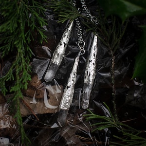 May include: Three silver pendant necklaces with clear quartz crystals hanging from chains. The necklaces are lying on a bed of green pine needles and brown leaves.