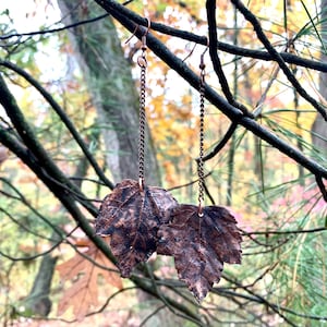 May include: A pair of dangle earrings featuring two copper leaf charms hanging from delicate chains. The earrings are photographed against a blurred background of fall foliage.