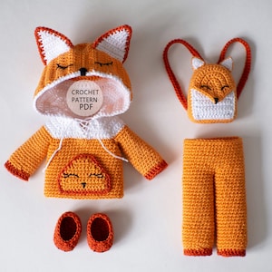 May include: A crocheted fox outfit for a doll or small toy. The outfit includes a hooded sweater, trousers, a backpack, and shoes. The sweater is orange and white with a fox face on the front. The trousers are orange with a red trim. The backpack is orange and white with a fox face on the front. The shoes are red.