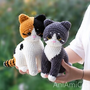 May include: Two handmade crochet cat plushies. One is a calico cat with orange, black, and white yarn, and the other is a grey and white cat. Both feature large, dark, glossy eyes, small pink noses, and white chest patches with textured yarn.