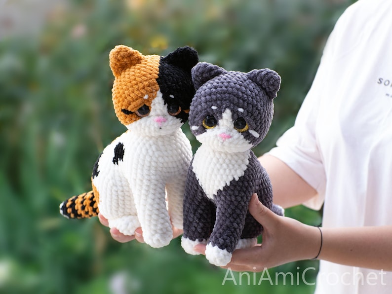 May include: Two handmade crochet cat plushies. One is a calico cat with orange, black, and white yarn, and the other is a grey and white cat. Both feature large, dark, glossy eyes, small pink noses, and white chest patches with textured yarn.