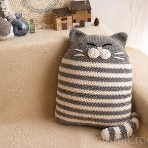 May include: A gray and white striped crocheted cat pillow with a white face and pink nose. The pillow is sitting on a light brown couch.