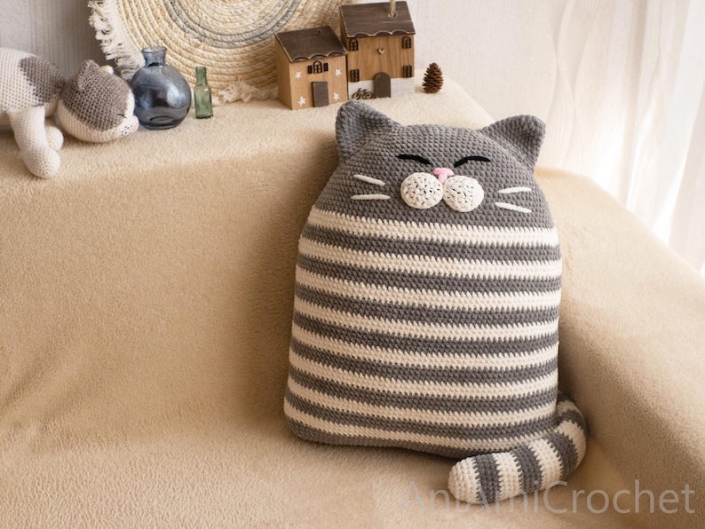 May include: A gray and white striped crocheted cat pillow with a white face and pink nose. The pillow is sitting on a light brown couch.