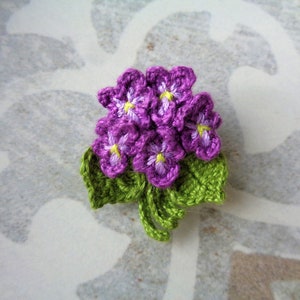 May include: A crocheted flower brooch with purple flowers and green leaves.