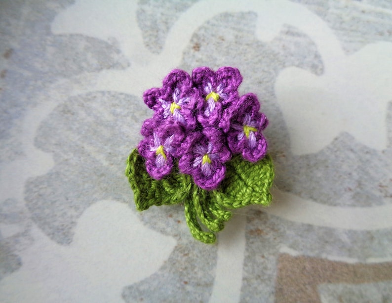 May include: A crocheted flower brooch with purple flowers and green leaves.