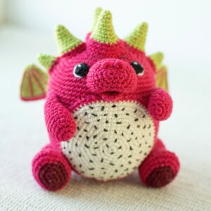 Op de afbeelding: Een gehaakt draken knuffel met een roze lichaam, groene stekels en een wit en zwart drakenfruit ontwerp op zijn buik.
