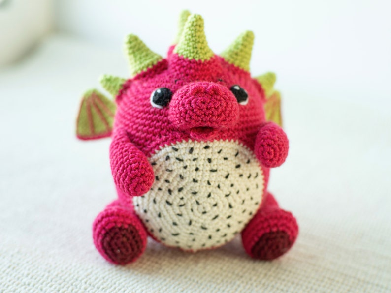 Op de afbeelding: Een gehaakt draken knuffel met een roze lichaam, groene stekels en een wit en zwart drakenfruit ontwerp op zijn buik.