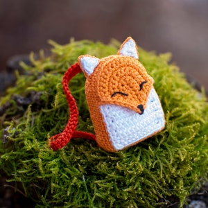 May include: A small crocheted fox backpack with orange and white fur, red straps, and a smiling face. The backpack is sitting on a bed of green moss.