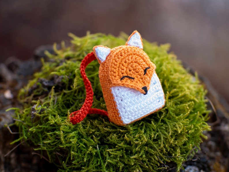 May include: A small crocheted fox backpack with orange and white fur, red straps, and a smiling face. The backpack is sitting on a bed of green moss.