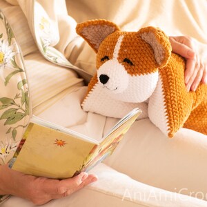 Peut inclure: Un jouet en peluche corgi crochet&eacute; &agrave; la main avec un motif en fil brun dor&eacute; et blanc, des oreilles pointues et un nez noir brod&eacute;. L'animal en peluche a une surface douce et textur&eacute;e et est pos&eacute; sur des genoux &agrave; c&ocirc;t&eacute; d'un livre ouvert.