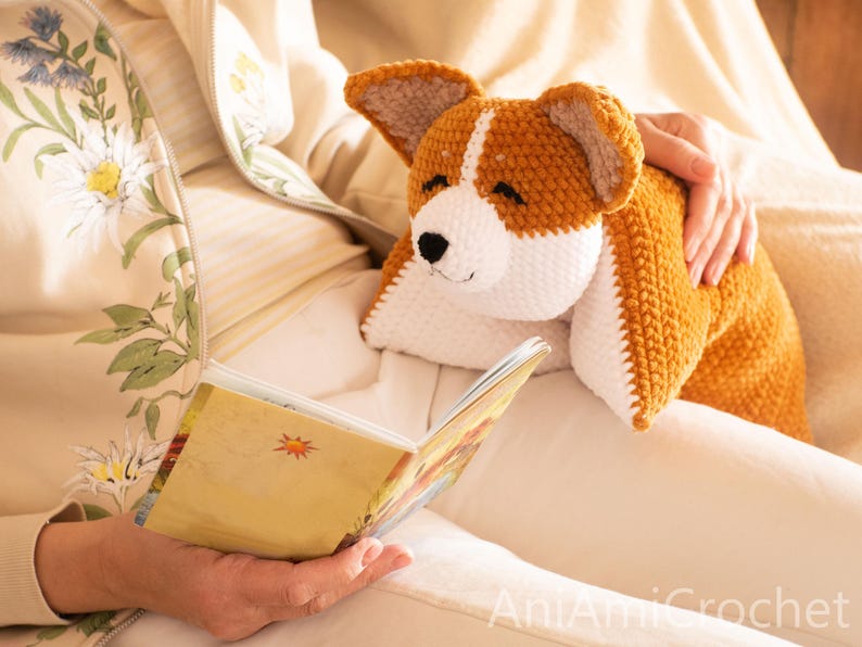 Peut inclure: Un jouet en peluche corgi crochet&eacute; &agrave; la main avec un motif en fil brun dor&eacute; et blanc, des oreilles pointues et un nez noir brod&eacute;. L'animal en peluche a une surface douce et textur&eacute;e et est pos&eacute; sur des genoux &agrave; c&ocirc;t&eacute; d'un livre ouvert.