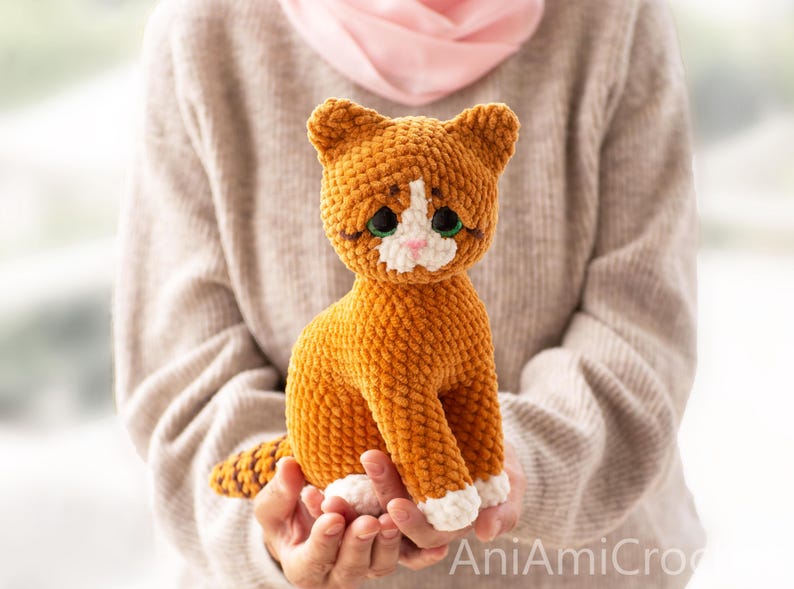 May include: A crocheted cat plush toy with orange fur, a white chest, white paws, and a white muzzle. The cat has large, dark green eyes with black pupils, a small pink nose, and stitched whiskers. The text AniAmiCro is visible in the bottom right corner.