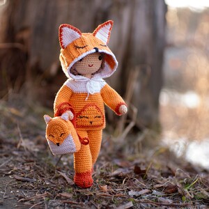 May include: A crocheted fox doll wearing an orange and white hooded sweater and carrying a small crocheted fox backpack. The doll is standing on a bed of brown leaves.