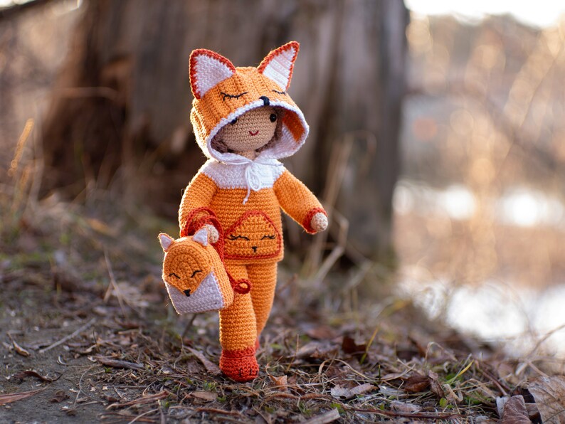 May include: A crocheted fox doll wearing an orange and white hooded sweater and carrying a small crocheted fox backpack. The doll is standing on a bed of brown leaves.