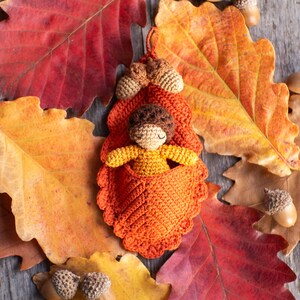 May include: A crocheted doll with brown hair and a yellow dress is sleeping in a crocheted orange leaf bed. The bed is surrounded by autumn leaves.