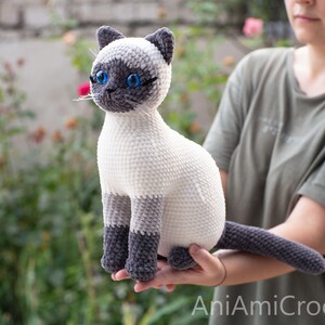 May include: A crocheted Siamese cat plush toy featuring a cream-colored body, dark gray ears, face mask, paws, and tail. The stuffed animal has bright blue plastic eyes, stitched whiskers, and a textured yarn finish. The text AniAmiCrochet is visible.