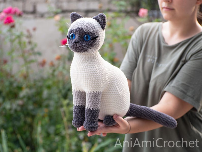 May include: A crocheted Siamese cat plush toy featuring a cream-colored body, dark gray ears, face mask, paws, and tail. The stuffed animal has bright blue plastic eyes, stitched whiskers, and a textured yarn finish. The text AniAmiCrochet is visible.