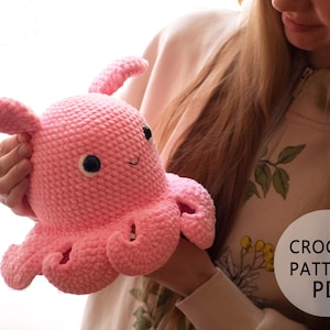 Puede incluir: Un juguete de amigurumi de pulpo de crochet rosa con ojos negros y una boca sonriente. El juguete está siendo sostenido por una persona. El texto "CROCHET PATTERN PDF" es visible en la imagen.