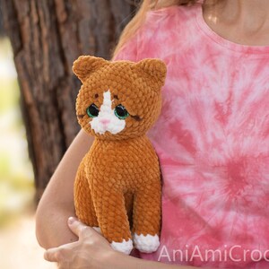 May include: A plush cat toy made of orange and white yarn with a soft, textured finish. The cat features large, dark eyes with green irises, a small pink nose, and white paws. The text AniAmiCrochet is visible in the bottom right corner.
