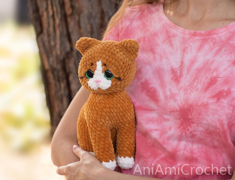 May include: A plush cat toy made of orange and white yarn with a soft, textured finish. The cat features large, dark eyes with green irises, a small pink nose, and white paws. The text AniAmiCrochet is visible in the bottom right corner.