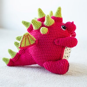 Op de afbeelding: Een roze gehaakte draak met groene stekels en een groene gehaakte vleugel. De draak heeft zwarte ogen en een witte gehaakte buik.