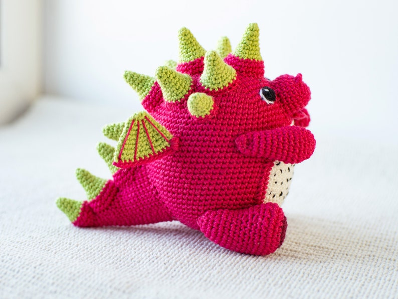 Op de afbeelding: Een roze gehaakte draak met groene stekels en een groene gehaakte vleugel. De draak heeft zwarte ogen en een witte gehaakte buik.