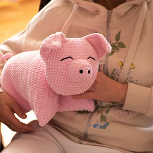 K&ouml;nnte beinhalten: Ein handgefertigtes hellrosa Pl&uuml;schschwein aus strukturiertem Garn. Das Schwein hat zwei dreieckige Ohren, eine abgerundete Schnauze mit zwei schwarzen gestickten Nasenl&ouml;chern und zwei geschlossene Augen, die durch einfache schwarze Linien dargestellt sind.
