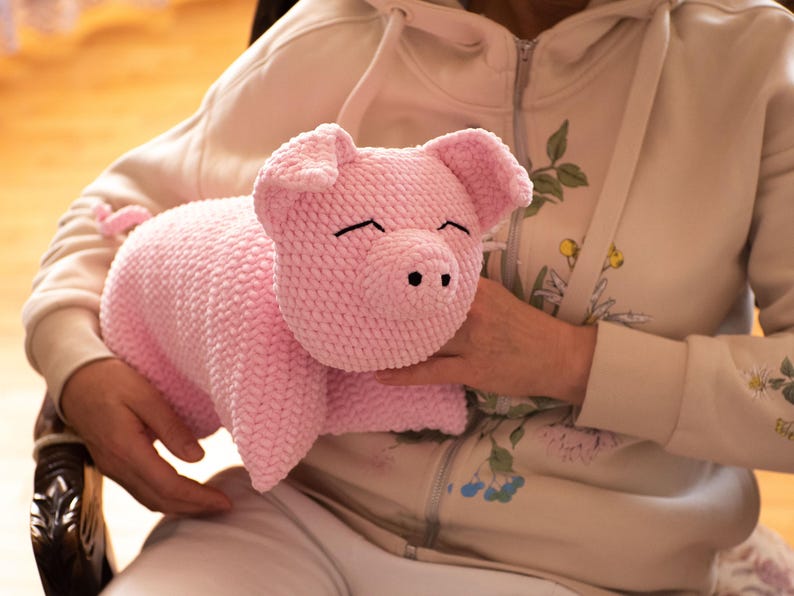 K&ouml;nnte beinhalten: Ein handgefertigtes hellrosa Pl&uuml;schschwein aus strukturiertem Garn. Das Schwein hat zwei dreieckige Ohren, eine abgerundete Schnauze mit zwei schwarzen gestickten Nasenl&ouml;chern und zwei geschlossene Augen, die durch einfache schwarze Linien dargestellt sind.