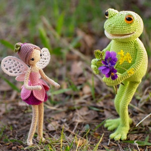 May include: A crocheted fairy doll with pink and white wings and a pink dress, standing next to a crocheted green frog holding a bouquet of purple and yellow flowers.