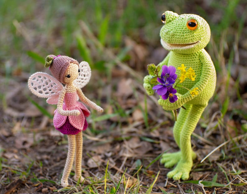 May include: A crocheted fairy doll with pink and white wings and a pink dress, standing next to a crocheted green frog holding a bouquet of purple and yellow flowers.