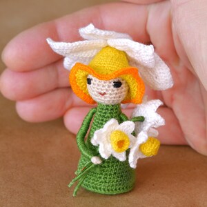 May include: A crocheted doll wearing a white flower crown with yellow and orange accents. The doll is holding a bouquet of white flowers with yellow centers. The doll is wearing a green dress.