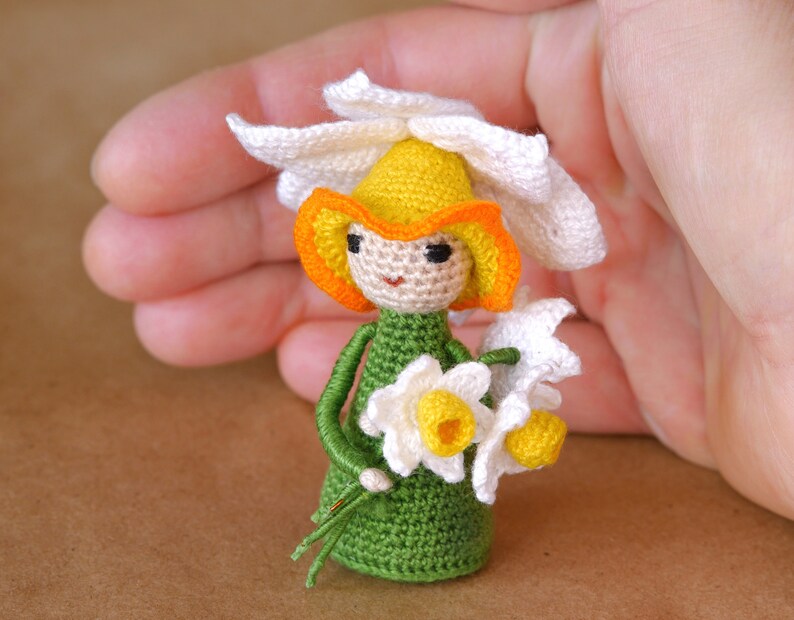 May include: A crocheted doll wearing a white flower crown with yellow and orange accents. The doll is holding a bouquet of white flowers with yellow centers. The doll is wearing a green dress.